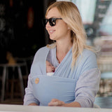 Woman wearing sunglasses and a light blue baby carrier with a baby inside, sitting at a table.